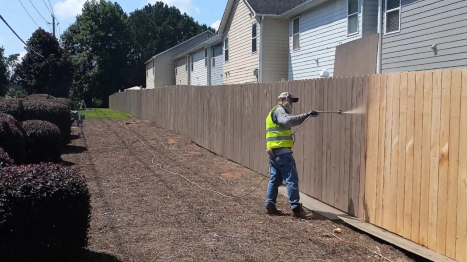 Wheeler Painting crew member in a safety vest staining a long wood privacy fence