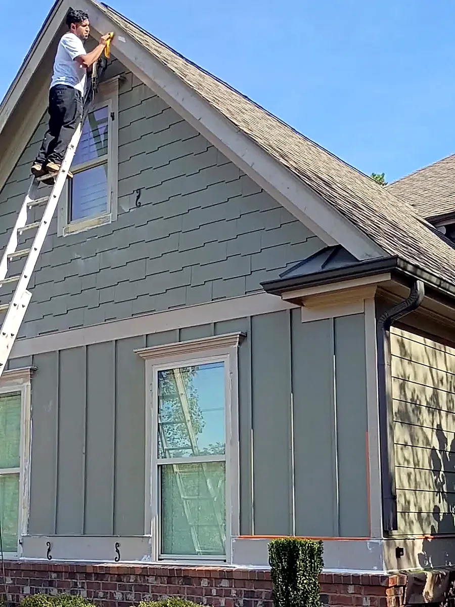 Wheeler Painting crew member on a ladder painting exterior trim on a green-sided Buford, GA home with masked windows